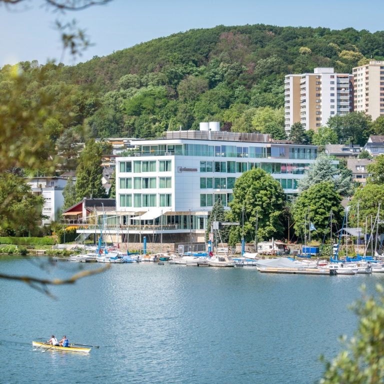 Modernes Hotel am Wasser mit Bootsanleger und grüner Hanglandschaft im Hintergrund.