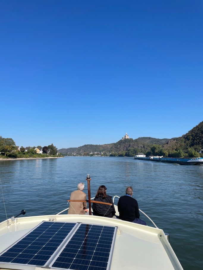 Drei Personen auf einem Boot, Blick auf den Fluss und eine hügelige Landschaft im Hintergrund.