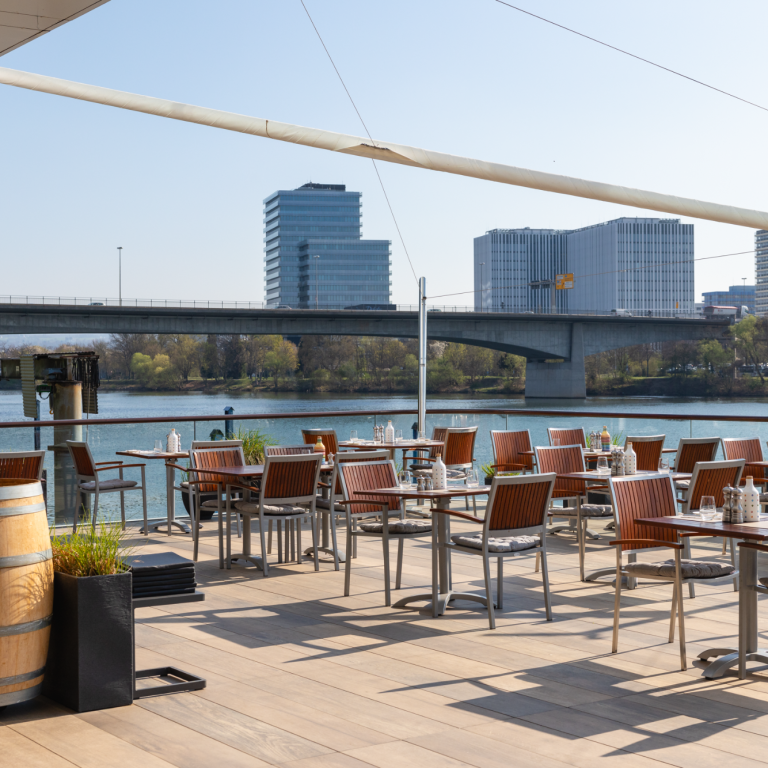 Terrasse mit Tischen am Wasser, Blick auf eine Stadtbrücke und moderne Gebäude im Hintergrund.