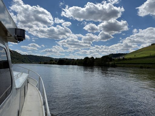 Blick auf einen Fluss mit Boot, grünen Hügeln und bewölktem Himmel im Hintergrund.