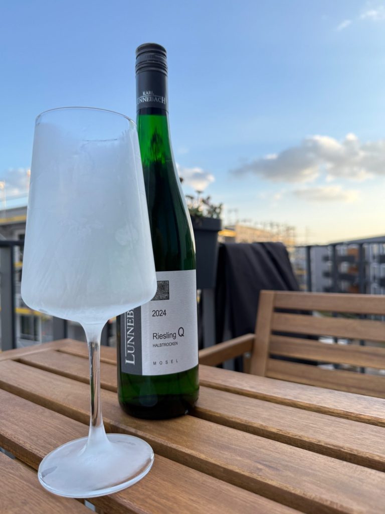 Ein gefrostetes Weinglas steht vor einer Flasche Riesling auf einem Holzstapel im Freien.