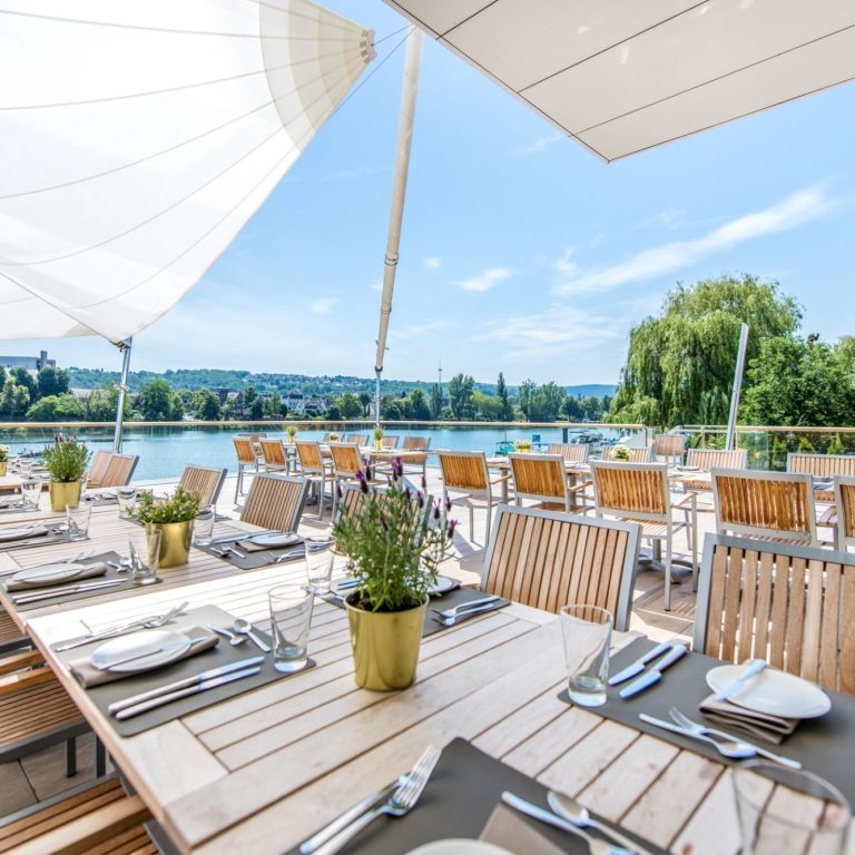 Terrasse mit Tischen am Wasser, Blick auf eine Stadtbrücke und moderne Gebäude im Hintergrund.